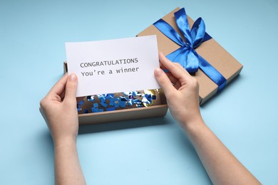 Photo of Woman holding card with phrase Congratulations You`re Winner and gift box on light blue background, closeup