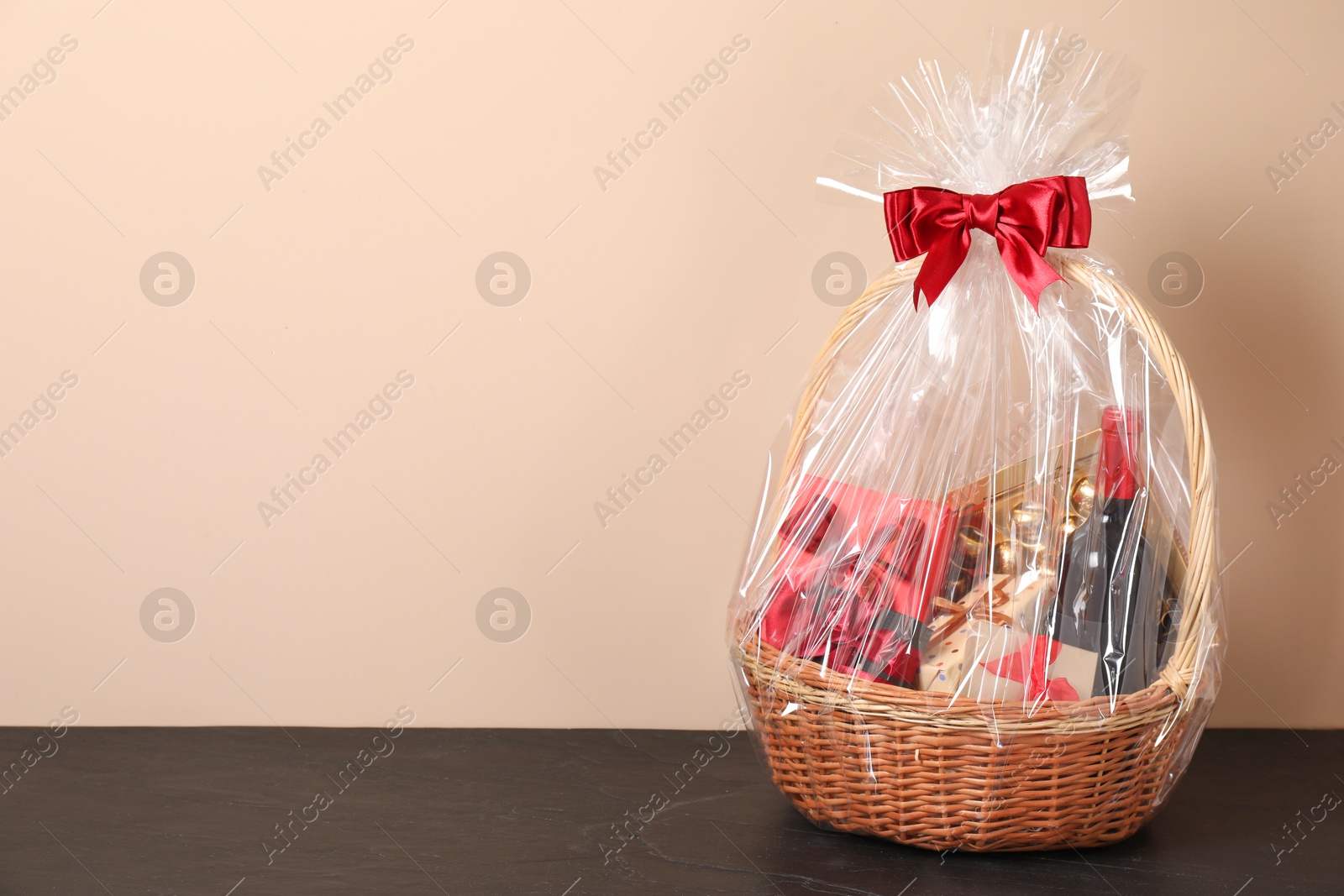 Photo of Wicker gift basket with wine on black textured table against beige background, space for text