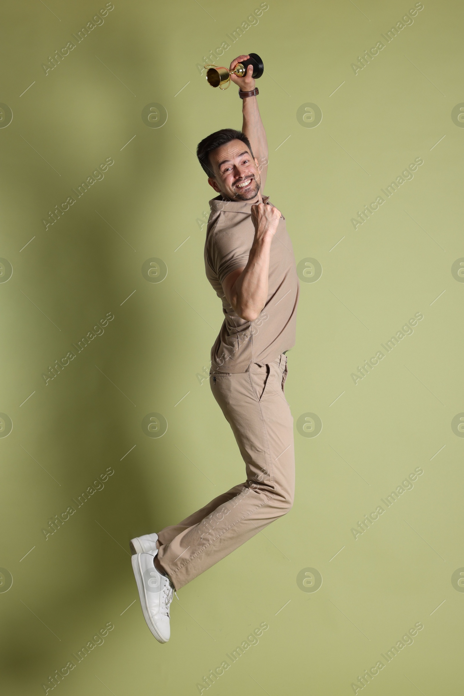 Photo of Happy winner with golden trophy cup jumping on pale olive background