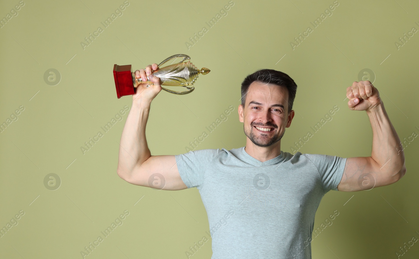 Photo of Happy winner with golden trophy cup showing his biceps on pale olive background