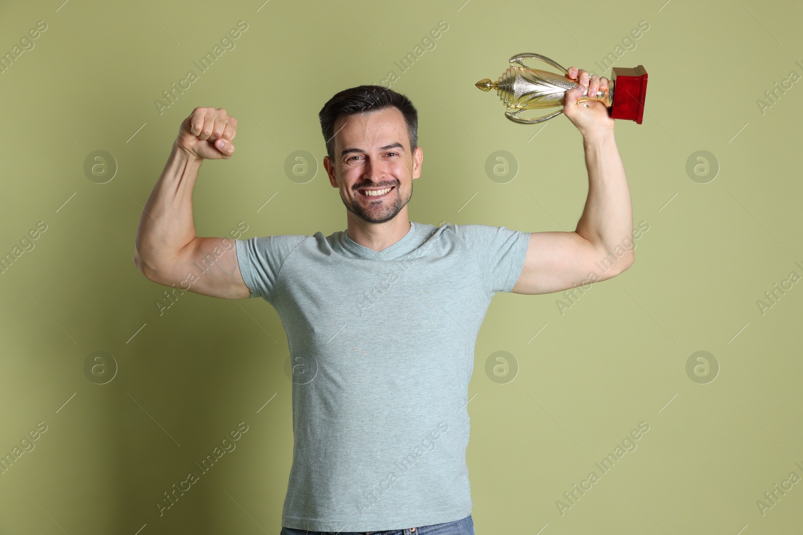 Photo of Happy winner with golden trophy cup showing his biceps on pale olive background