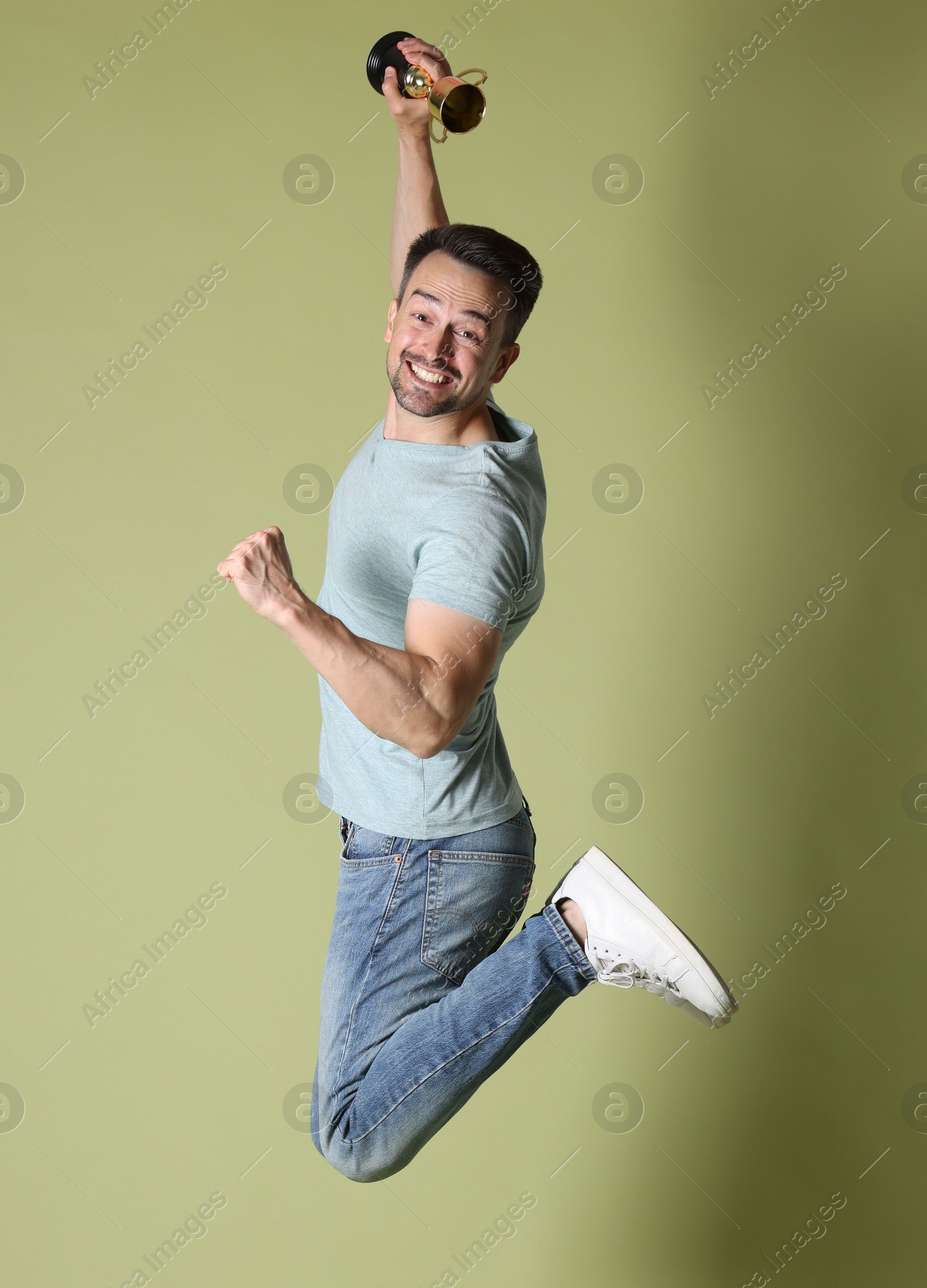 Photo of Happy winner with golden trophy cup jumping on pale olive background