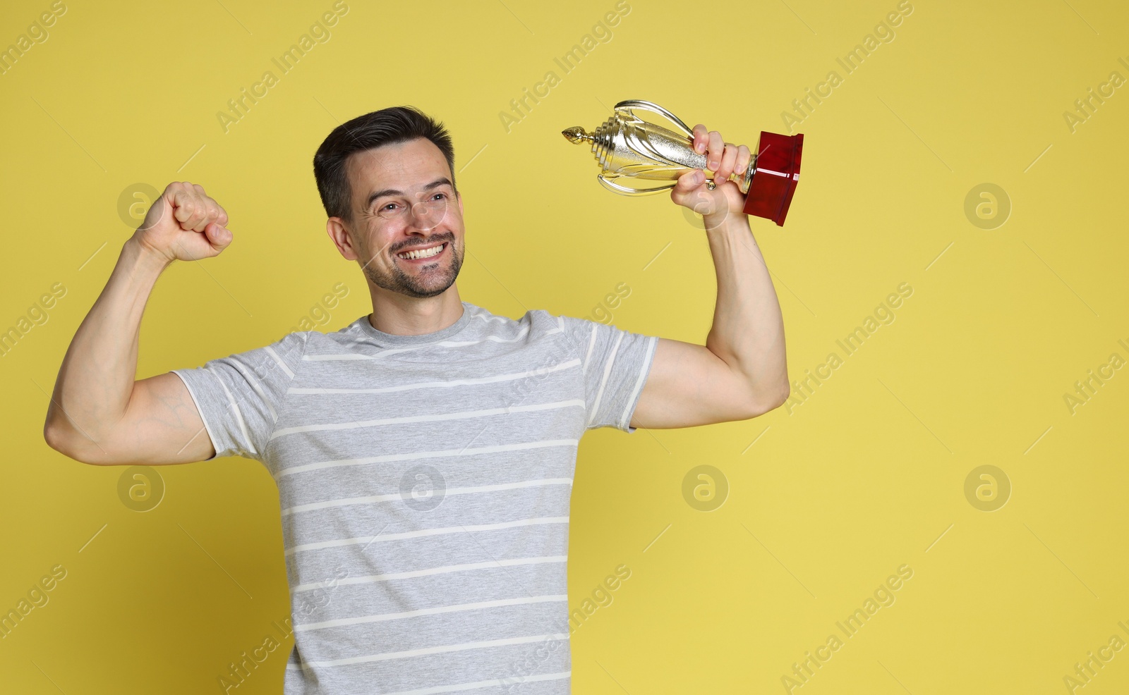 Photo of Happy winner with golden trophy cup showing his biceps on yellow background