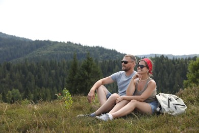 Photo of Happy couple with backpack resting on grass outdoors, space for text. Active tourism