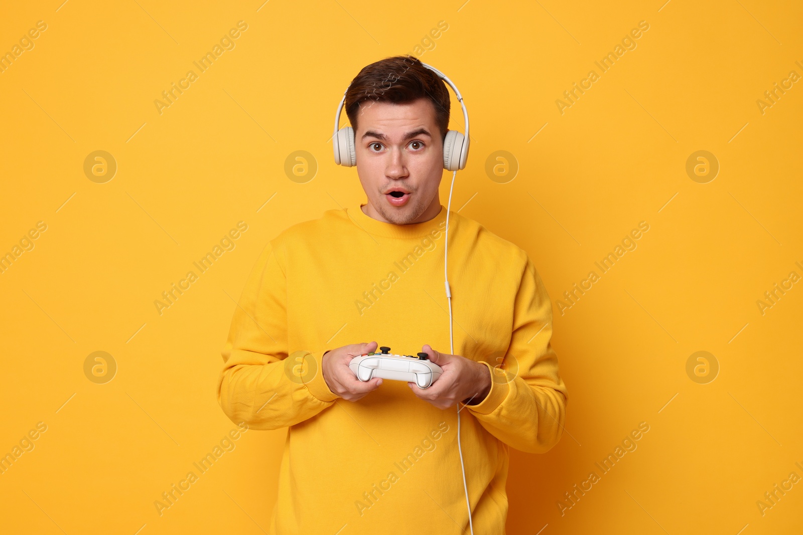 Photo of Young man in headphones playing video games with controller on orange background