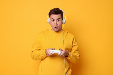 Young man in headphones playing video games with controller on orange background