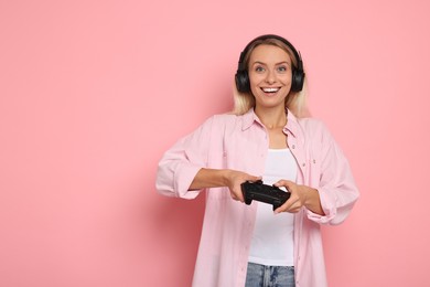 Happy woman in headphones playing video games with controller on pink background, space for text