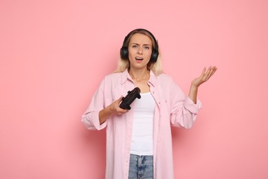 Unhappy woman in headphones playing video games with controller on pink background