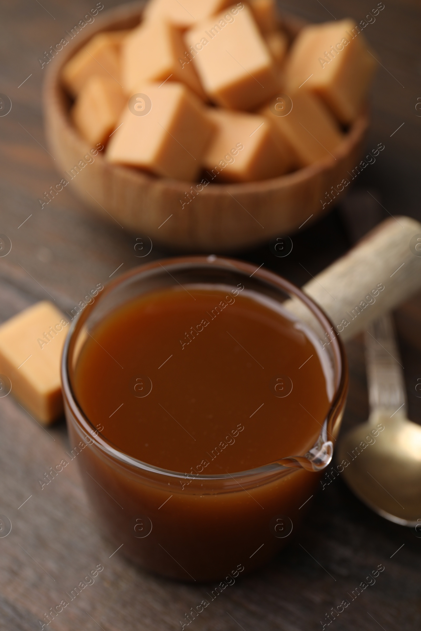 Photo of Tasty caramel sauce in glass measuring cup and sweet candies on wooden table, closeup