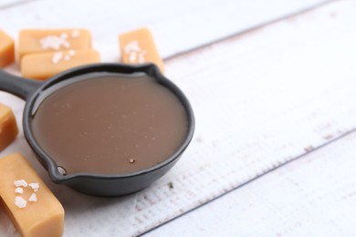 Photo of Tasty salted caramel in gravy boat and candies on white wooden table, closeup. Space for text