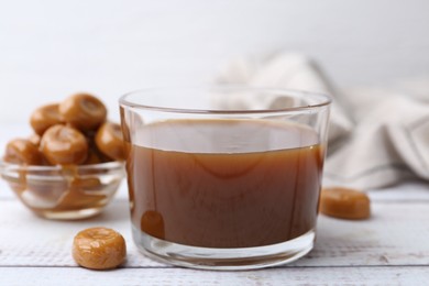 Photo of Tasty caramel sauce in bowl and candies on white wooden table, closeup