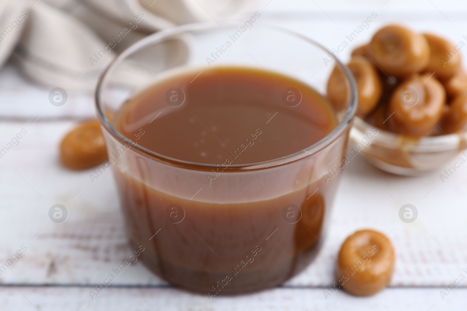 Photo of Tasty caramel sauce in bowl and candies on white wooden table, closeup