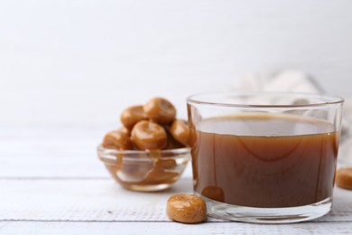 Photo of Tasty caramel sauce in bowl and candies on white wooden table, closeup