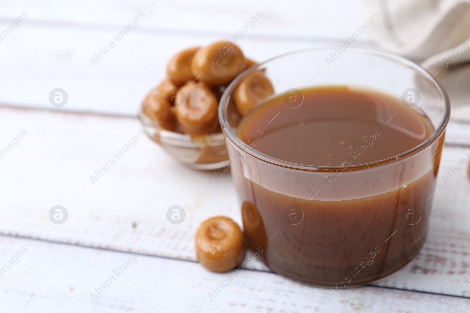 Photo of Tasty caramel sauce in bowl and candies on white wooden table, closeup. Space for text