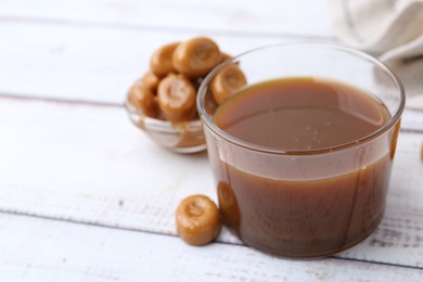 Photo of Tasty caramel sauce in bowl and candies on white wooden table, closeup. Space for text