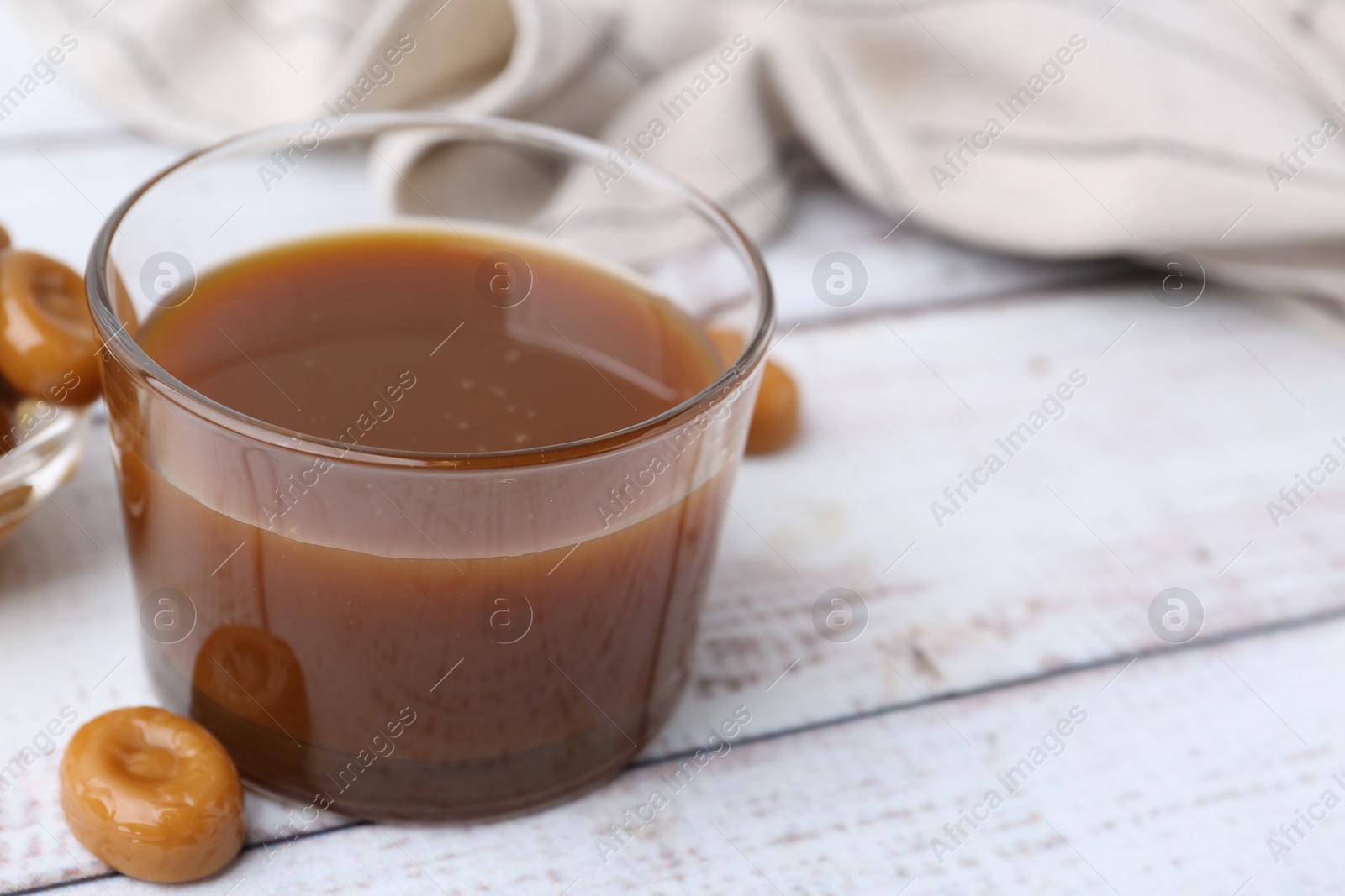 Photo of Tasty caramel sauce in bowl and candies on white wooden table, closeup. Space for text