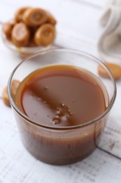 Tasty caramel sauce in bowl and candies on white wooden table, closeup