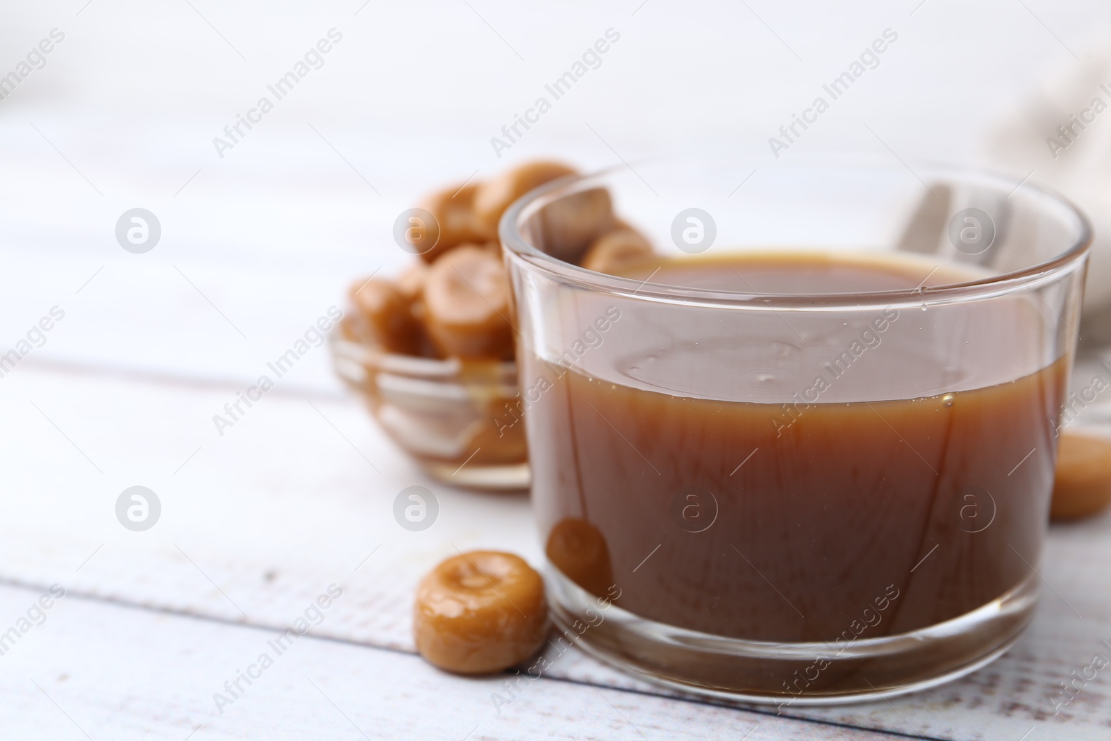 Photo of Tasty caramel sauce in bowl and candies on white wooden table, closeup