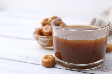 Photo of Tasty caramel sauce in bowl and candies on white wooden table, closeup