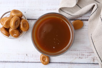 Photo of Tasty caramel sauce in bowl and candies on white wooden table, top view