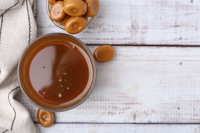 Photo of Tasty caramel sauce in bowl and candies on white wooden table, top view. Space for text