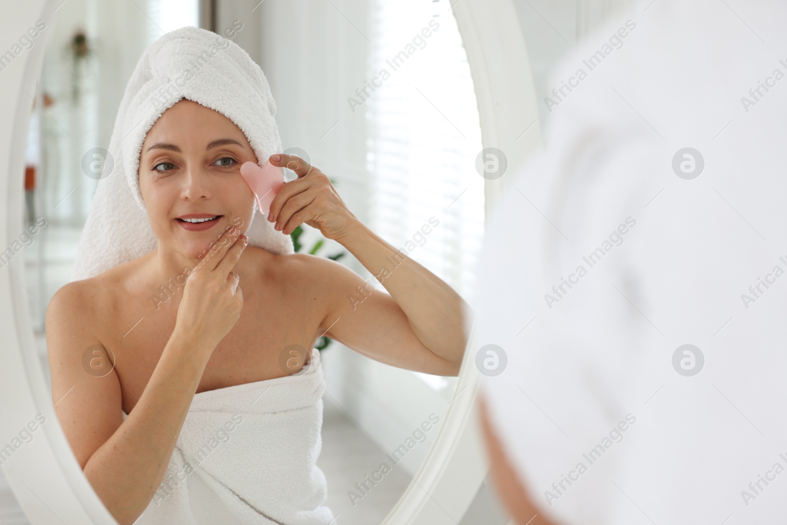 Photo of Smiling woman doing facial self massage with gua sha tool near mirror in bathroom