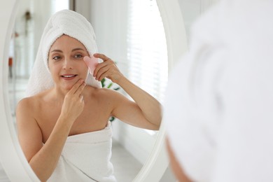 Smiling woman doing facial self massage with gua sha tool near mirror in bathroom