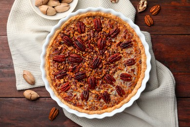 Photo of Delicious pecan pie in baking dish and fresh nuts on wooden table, flat lay