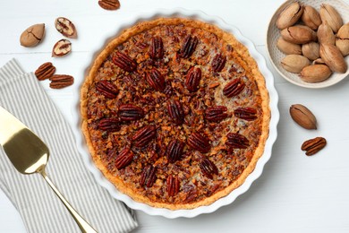 Photo of Delicious pecan pie in baking dish, fresh nuts and cake server on white wooden table, flat lay