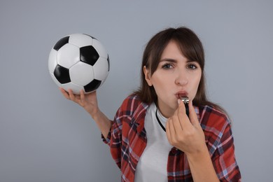 Woman with soccer ball blowing whistle on grey background