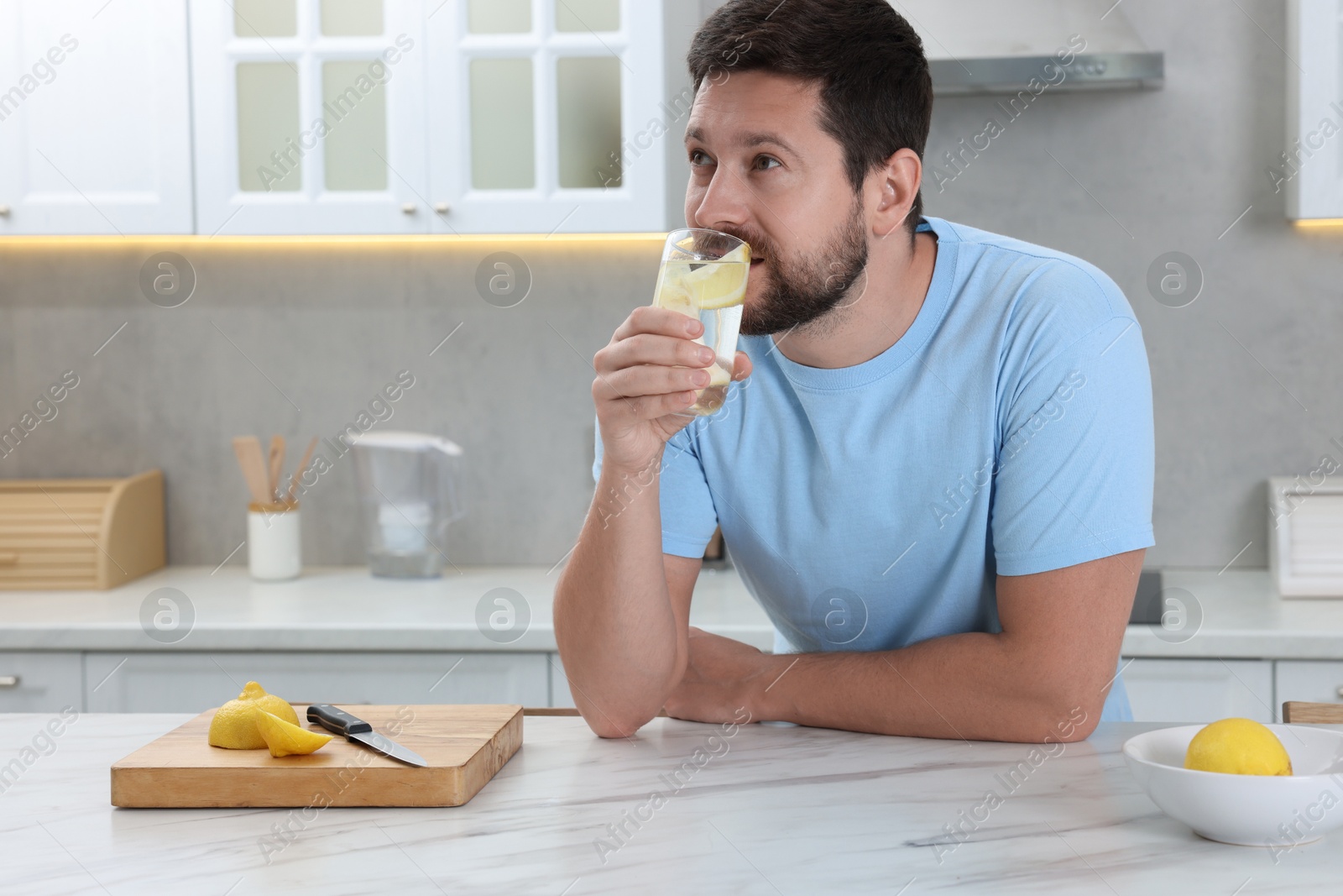 Photo of Handsome man drinking water with lemon at white marble table in kitchen