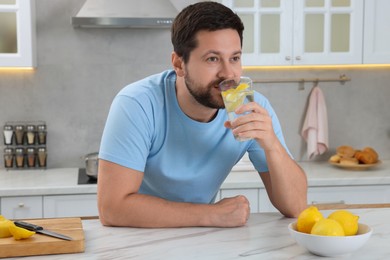 Photo of Handsome man drinking water with lemon at white marble table in kitchen