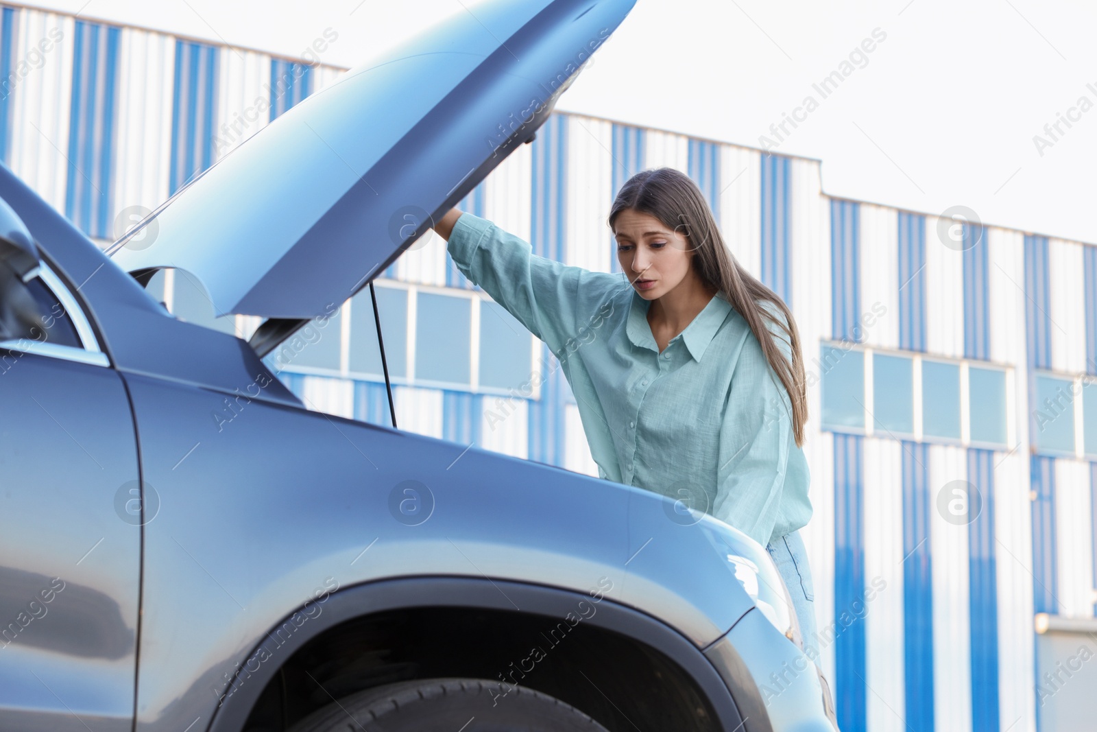 Photo of Stressed woman looking under hood of broken car outdoors