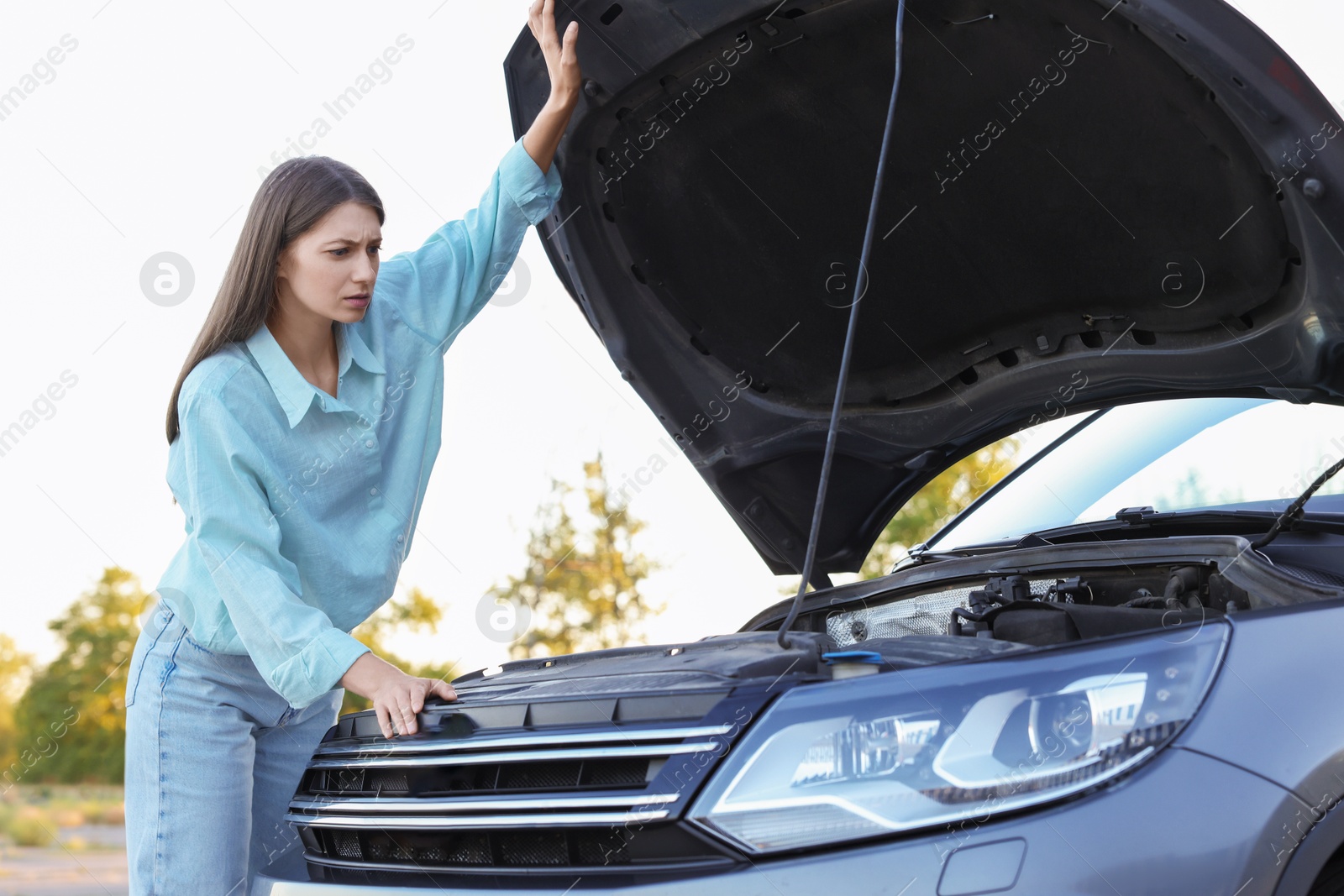 Photo of Stressed woman looking under hood of broken car outdoors