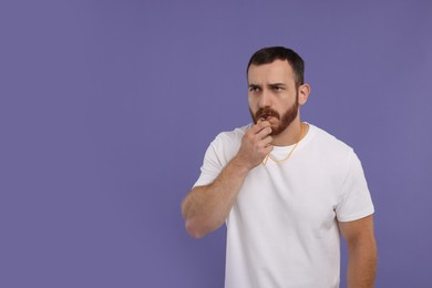 Young man blowing whistle on purple background, space for text