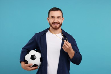 Happy young man with whistle and soccer ball on light blue background