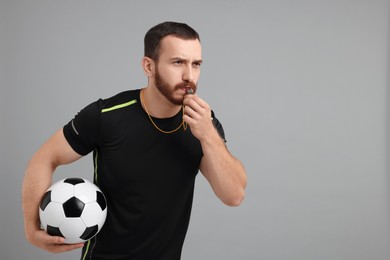 Young man with soccer ball blowing whistle on grey background, space for text