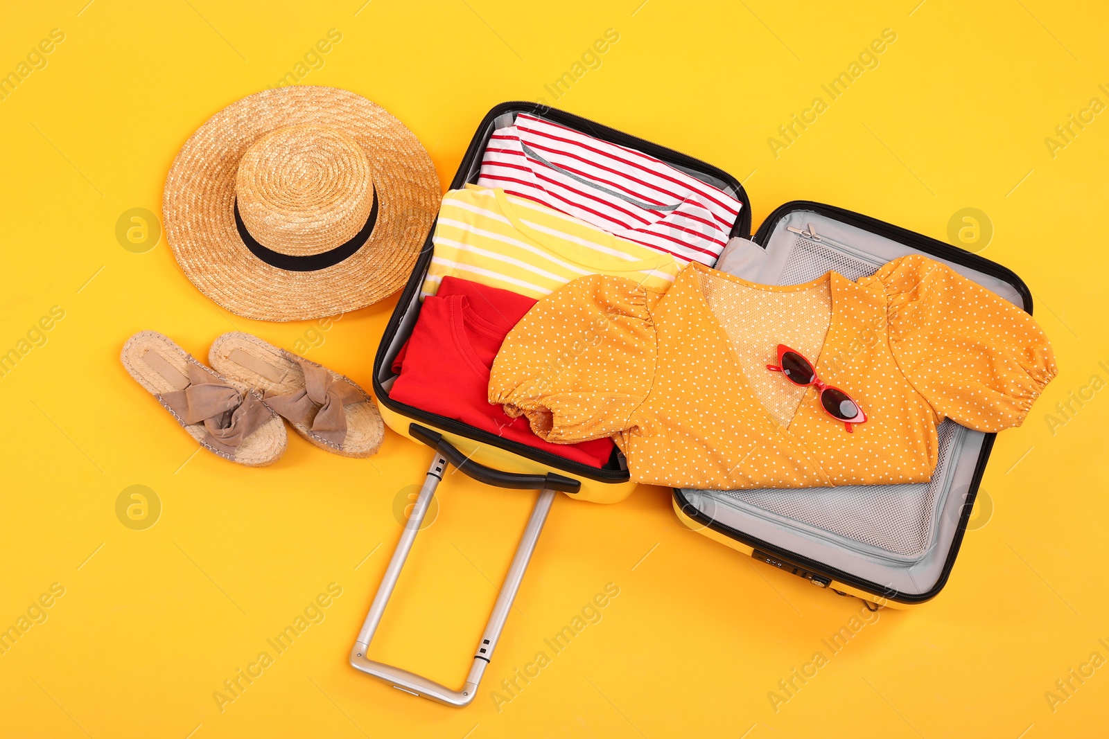 Photo of Open suitcase with clothes, wicker hat and shoes on yellow background, flat lay