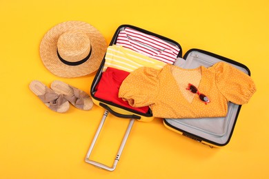 Photo of Open suitcase with clothes, wicker hat and shoes on yellow background, flat lay