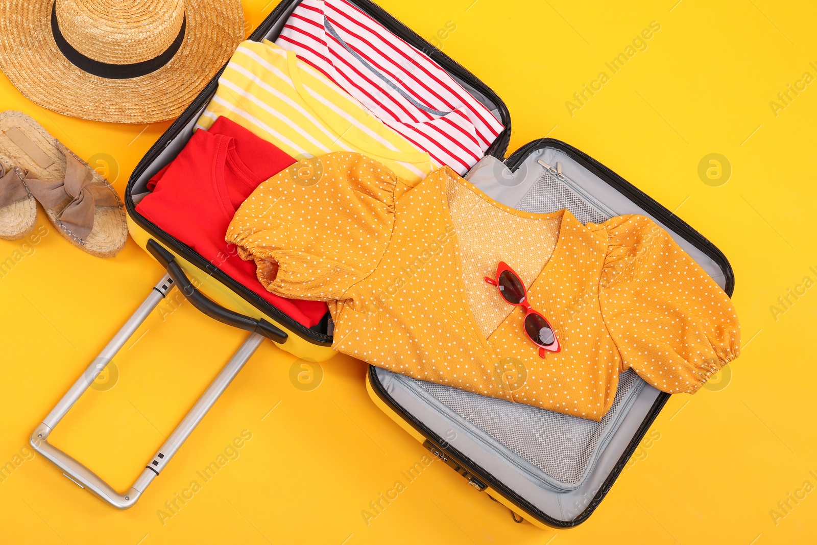 Photo of Open suitcase with clothes, wicker hat and shoes on yellow background, flat lay