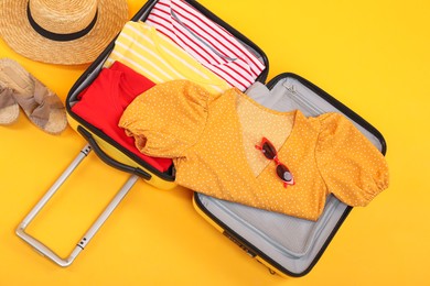 Photo of Open suitcase with clothes, wicker hat and shoes on yellow background, flat lay