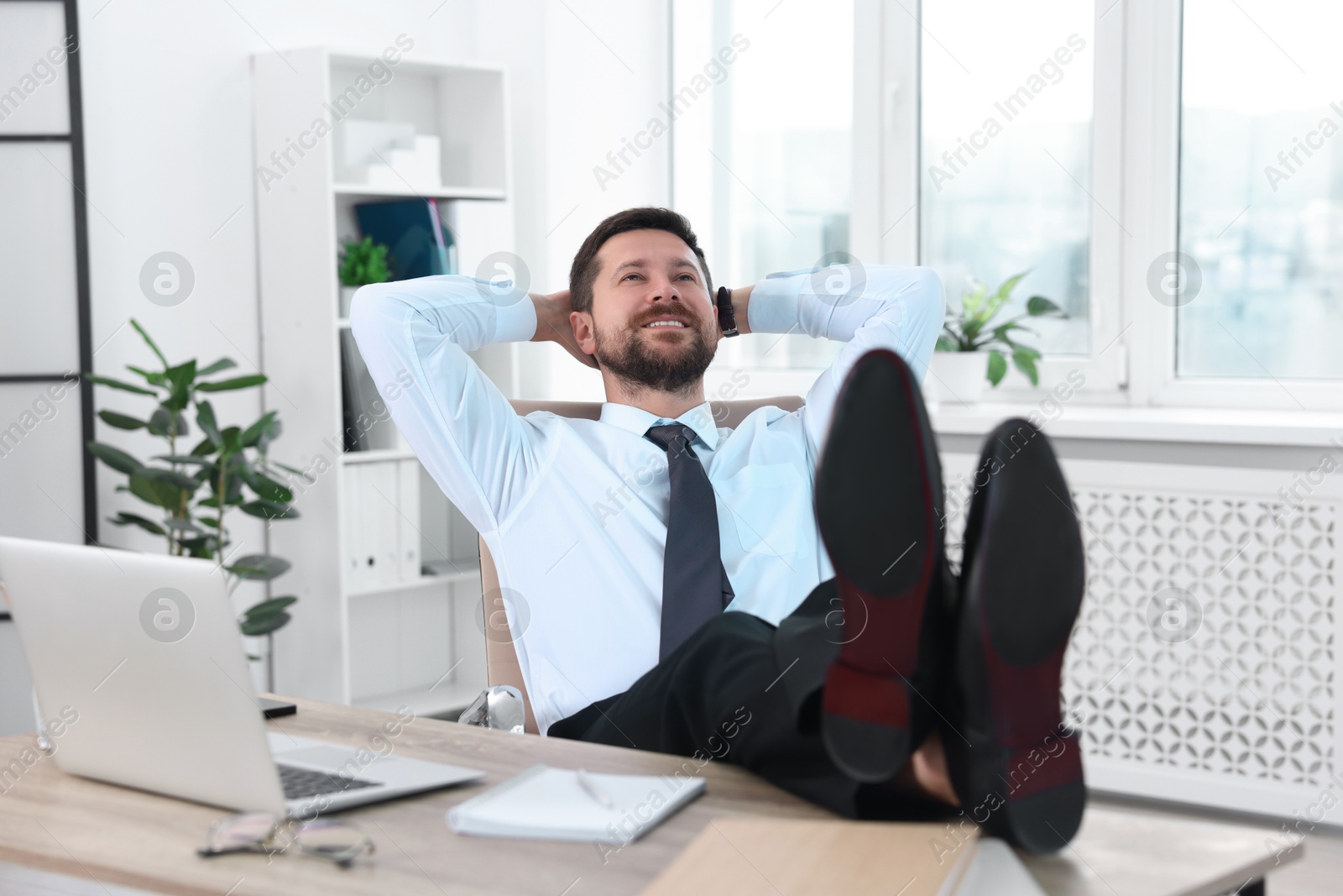 Photo of Smiling businessman with hands behind his head holding legs on table in office. Break time