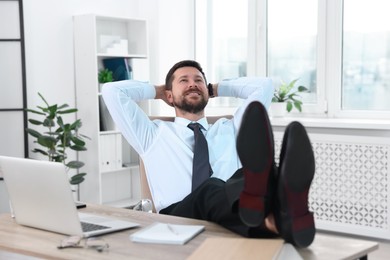 Photo of Smiling businessman with hands behind his head holding legs on table in office. Break time