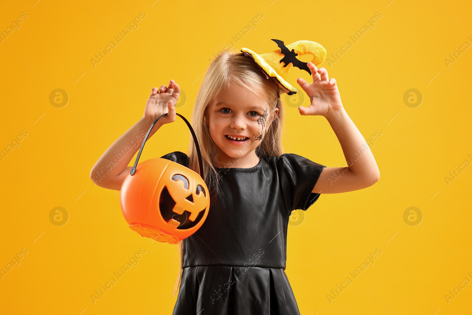 Photo of Cute girl with pumpkin bucket dressed like witch on yellow background. Halloween celebration