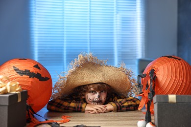 Cute boy dressed like scarecrow with festive decor and gift boxes indoors at night. Halloween celebration