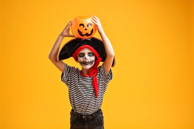 Photo of Funny boy with pumpkin bucket dressed like pirate on yellow background. Halloween costume