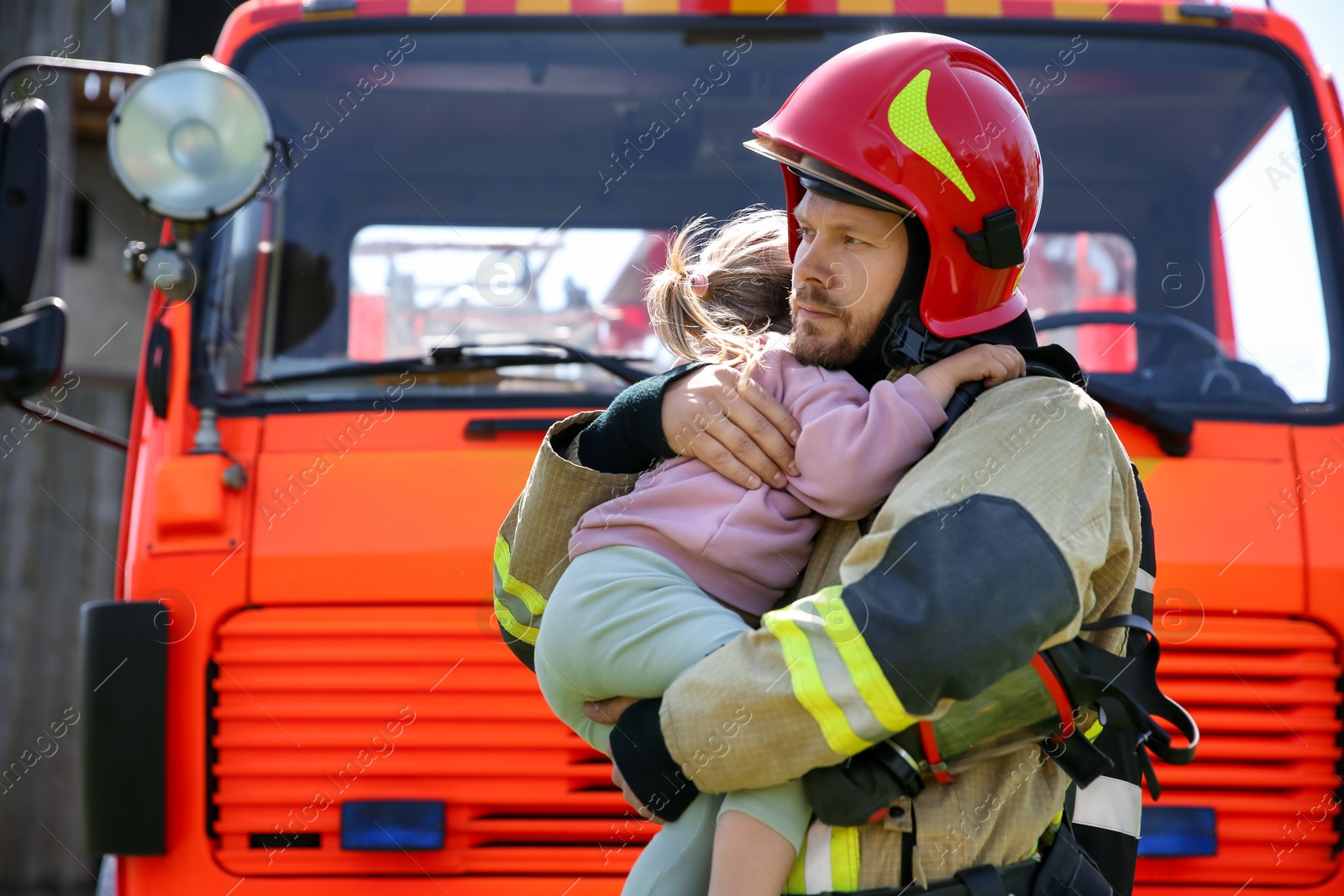 Photo of Firefighter in uniform holding rescued little girl near fire truck outdoors. Save life