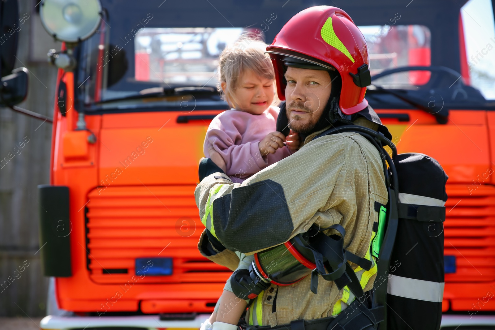 Photo of Firefighter in uniform holding rescued little girl near fire truck outdoors. Save life