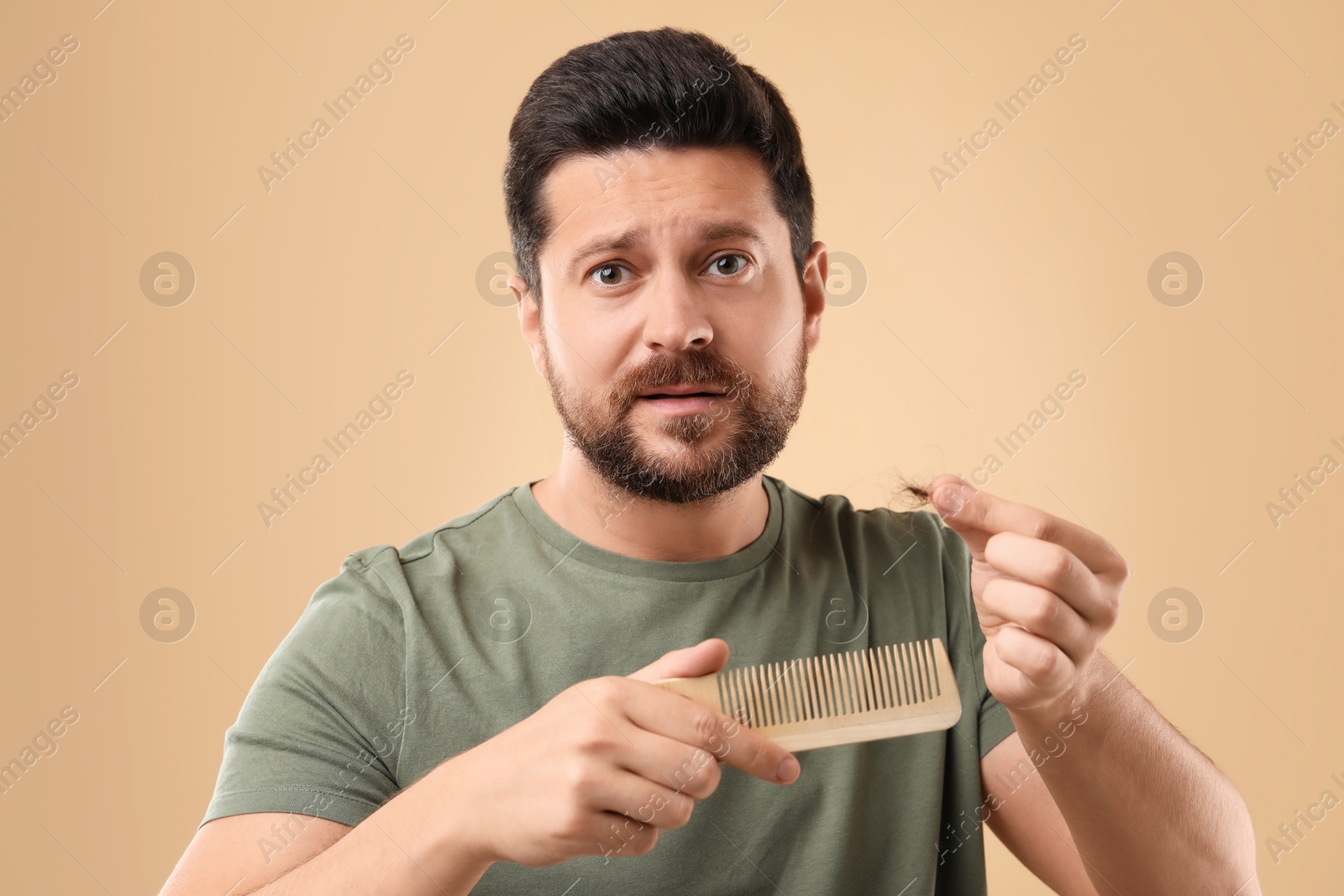 Photo of Emotional man holding clump of lost hair and comb on beige background. Alopecia problem