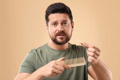 Photo of Emotional man holding clump of lost hair and comb on beige background. Alopecia problem